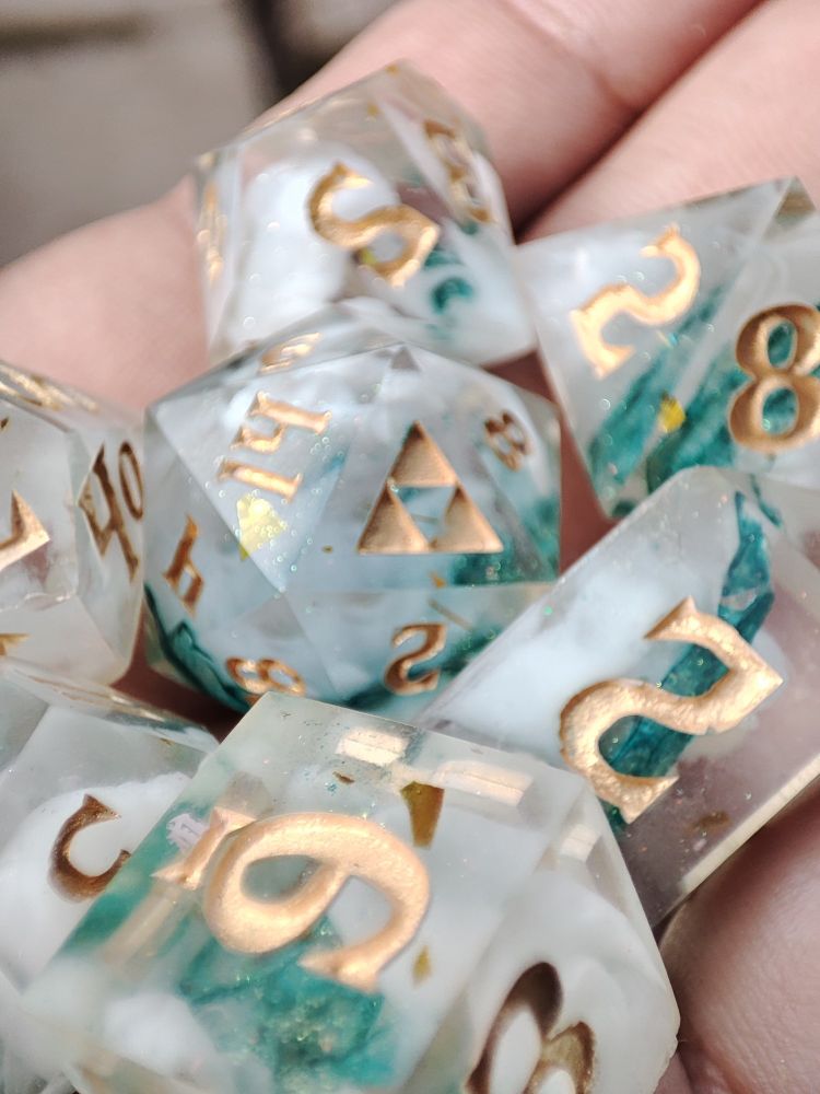 Most of a 7 piece set of dice inside someones hand. The d20 has a triforce on the 20 face, and inside the dice are opaque  white clouds and a strip of turquoise dyed snake shed. 