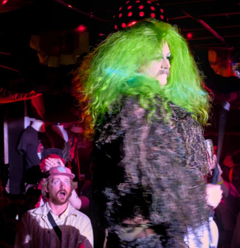 A drag queen in a neon green wig is performing in the foreground. In the background, a man in a red hat gasps.