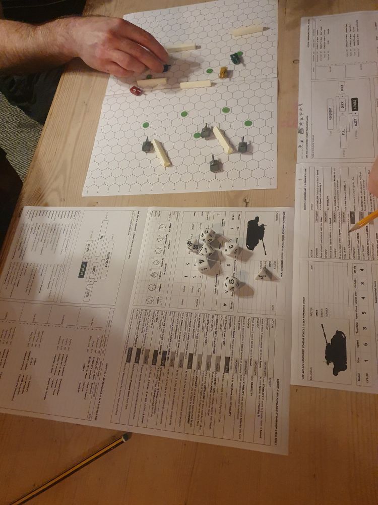 A photo of a tabletop role-playing game. One player is moving a tiny model of a tank around a hexagonal board. The board is surrounded by character sheets, dice and pencils.