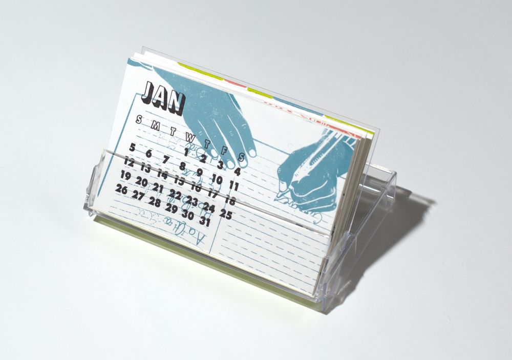 Small letterpress printed cards in a cassette tape case. The card shows the calendar days January 2025 in a grid with the weekdays with the word JAN printed above. The weekdays are abbreviated as S, M, T, W, T, F, S. A pair of blue hands writes into a school notebook.