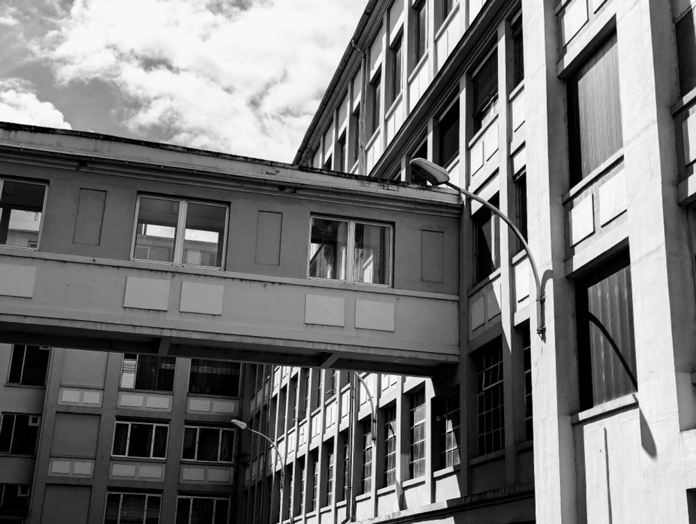 Ein alter, gefühlt aus den 70ern stammender Industriegebäude-Komplex ist schwarz-weiß zu sehen. Aus der Wand ragt eine Straßenlaterne heraus. Eine Brücke verbindet zwei Gebäude.