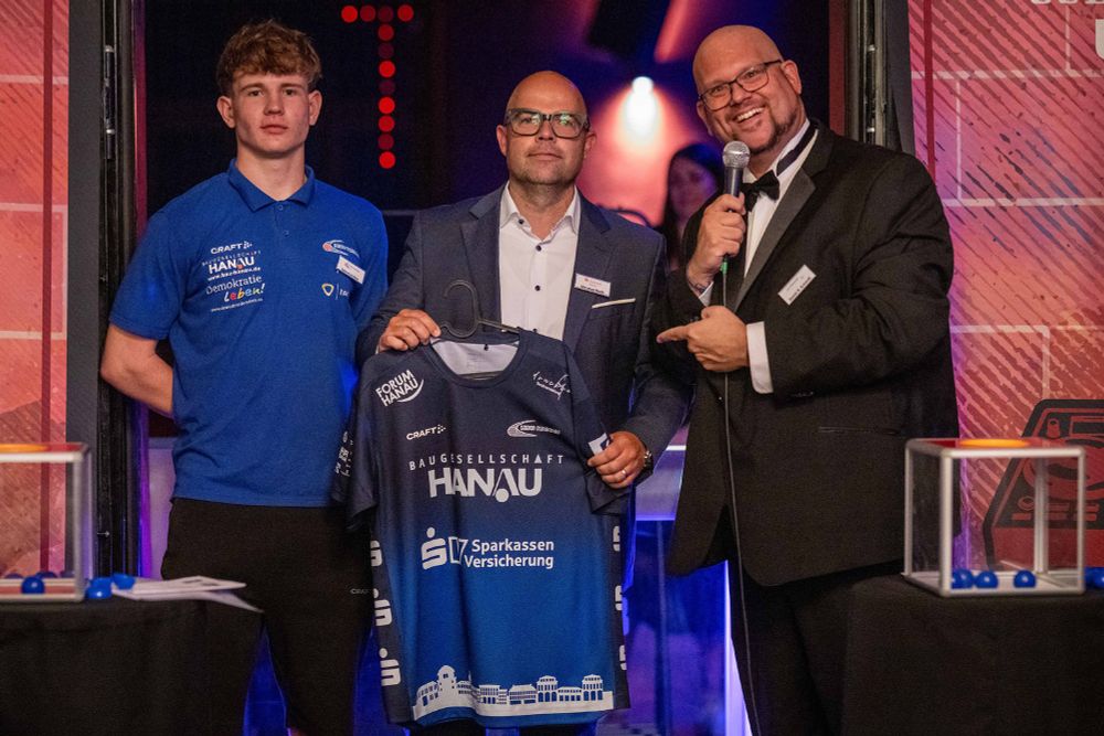 Christian Rieth von der Sparkasse Hanau präsentiert ein auf einem Bügel trapiertes Trikot, bei dem die Sparkassen-Versicherung präsent auf der Vorderseite zu sehen ist. Er wird eingerahmt vom HSG-Hanau-Spieler Sebastian Hein sowie Moderator Daniel R. Schmidt.