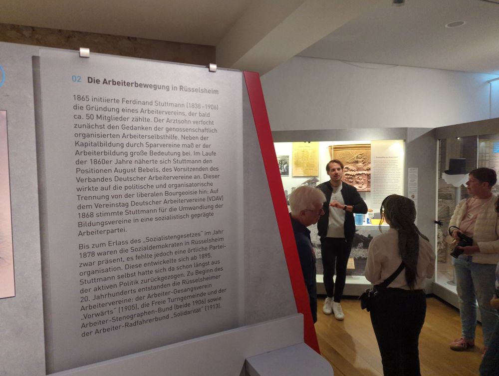 Ein Schild im Museum Rüsselsheim zeigt einen Informationstext über die Arbeiterbewegung. Im Hintergrund spricht der Museumsführer zu den Mitgliedern des Social Media Clubs Frankfurt.