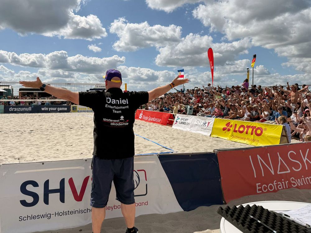 Daniel R. Schmidt steht an der Bande eines Volleyball-Courts und steht mit dem Rücken zur Kamera. Er steht dem Publikum auf der seitlichen Tribüne zugewandt und formt mit seinen nach oben gestreckten Armen ein V.
