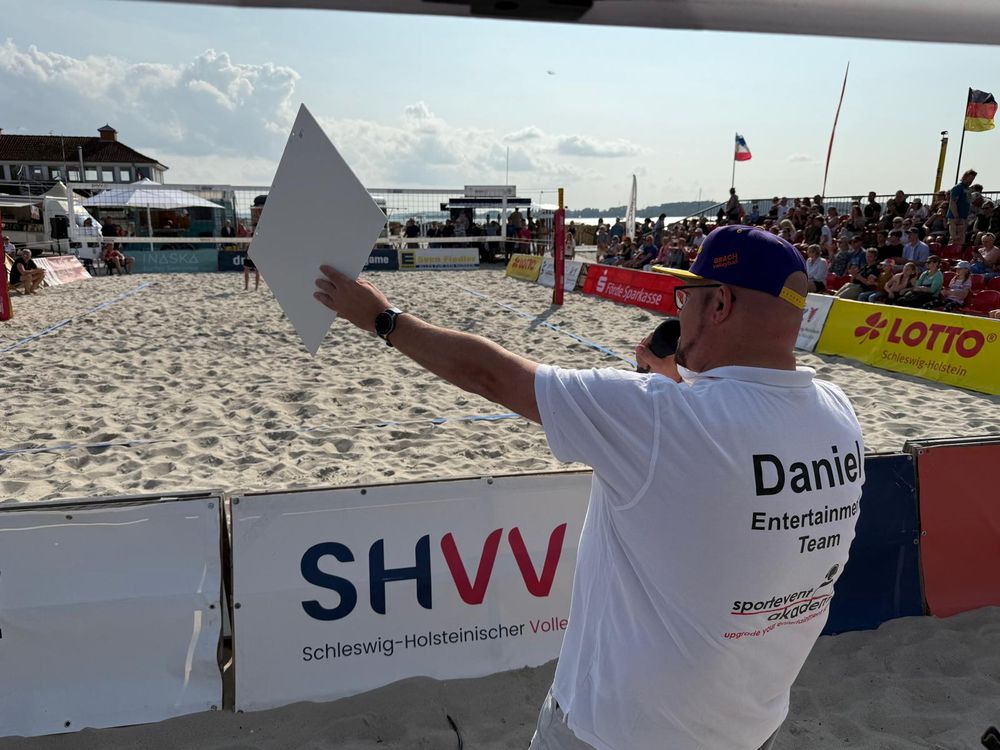 Daniel R. Schmidt steht mit dem Rücken zur Kamera und an der Bande eines Beach-Volleyball-Courts. Schräg gegenüber sitzen Zuschauer:innen und schauen auf eine Spielerin, die gerade beim Einlaufen ist, aber durch den Bildrand verdeckt ist. Daniel hält ein großes weißen Klemmbrett in die Richtung der Spielerin und spricht in sein Mikrofon.