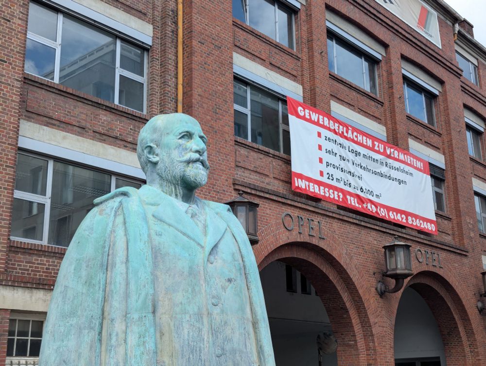 Eine Statue von Adam Opel steht vor dem Backsteinbau des Opel-Altwerks. Ein rot-weißes Werbebanner stört den architektonischen Eindruck erheblich.