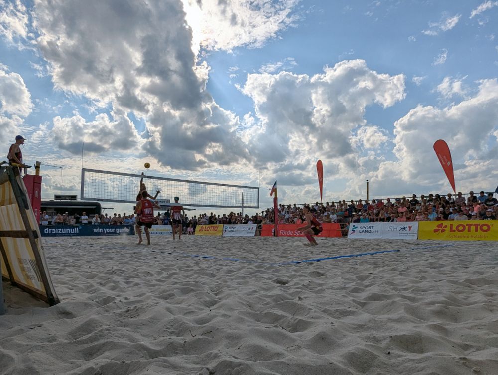 Das Foto zeigt einen Beach-Volleyball-Court auf der Höhe des Sandes. Ein Spieler springt am Netz gerade hoch und greift an. Auf der anderen Feldseite steht ein Blockspieler und ein Abwehrspieler in den Knien. Gegenüber steht eine vollbesetzte Zuschauer:innen-Tribüne.