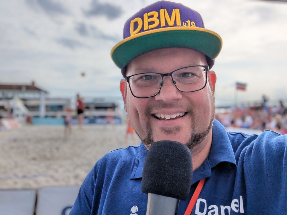 Daniel R. Schmidt schießt mit Mikrofon in der Hand ein Selfie, bei dem im Hintergrund eine Spielszene eines Beach-Volleyball-Courts unscharf im Hintergrund zu sehen ist.