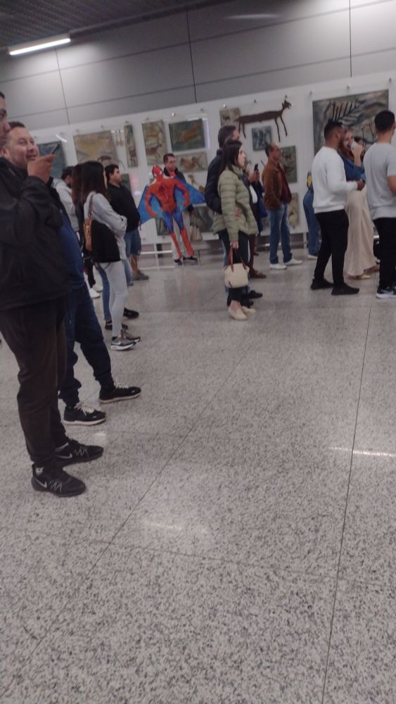 Grupo de pessoas parado em frente ao lugar de  desembarque do aeroporto. Ao fundo é possível ver uma pessoa segurando um homi aranha inflável e a parede está coberta por artes e quadros 