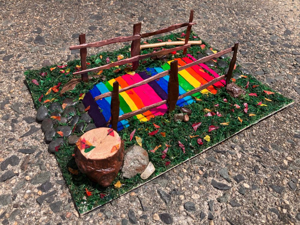One view of a 3D scene sculpture of a nature trail bridge. The bridge path is rainbow wooden planks (pink, red, orange, yellow, green, teal, blue, purple), there is grass coming up through some of the planks as well as leaves on some of them. And the poles and railing of the bridge are wooden and brown. On both sides of the bridge ends there are stone pebble paths with grass growing around them. There is grass around the sides of the bridge, there are leaves in the grass. In the corner in the grass there is a tree stump with a few leaves on it, there are 2 rocks against it. There are several rocks against the bridge poles as well. In another corner there is a wooden branch in the grass. 