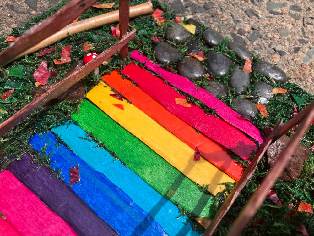 A close up view of a part of a 3D scene sculpture of a nature trail bridge. This close up features one pebble path at the end of the bridge. The bridge path is rainbow wooden planks (pink, red, orange, yellow, green, teal, blue, purple), there is grass coming up through some of the planks as well as leaves on some of them. And the poles and railing of the bridge are wooden and brown. At the bridge end there is a stone pebble path with grass growing around them. There are several rocks leaning against the bridge poles. There is a wooden branch in the corner in the grass. 