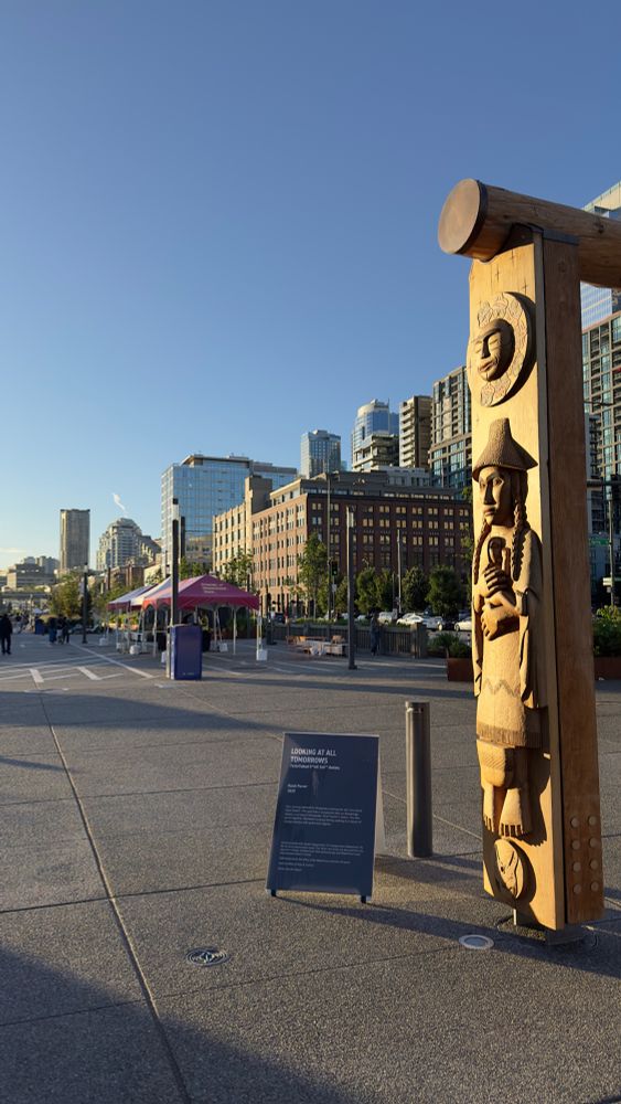 A Northwest native sculpture along next to a broad promenade with the Seattle skyline behind. Lovely sunset light. 