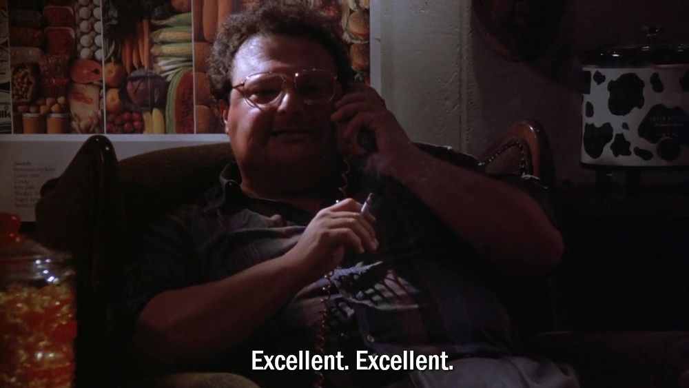 Newman from Seinfeld smoking a cigarette in a darkened room saying, 