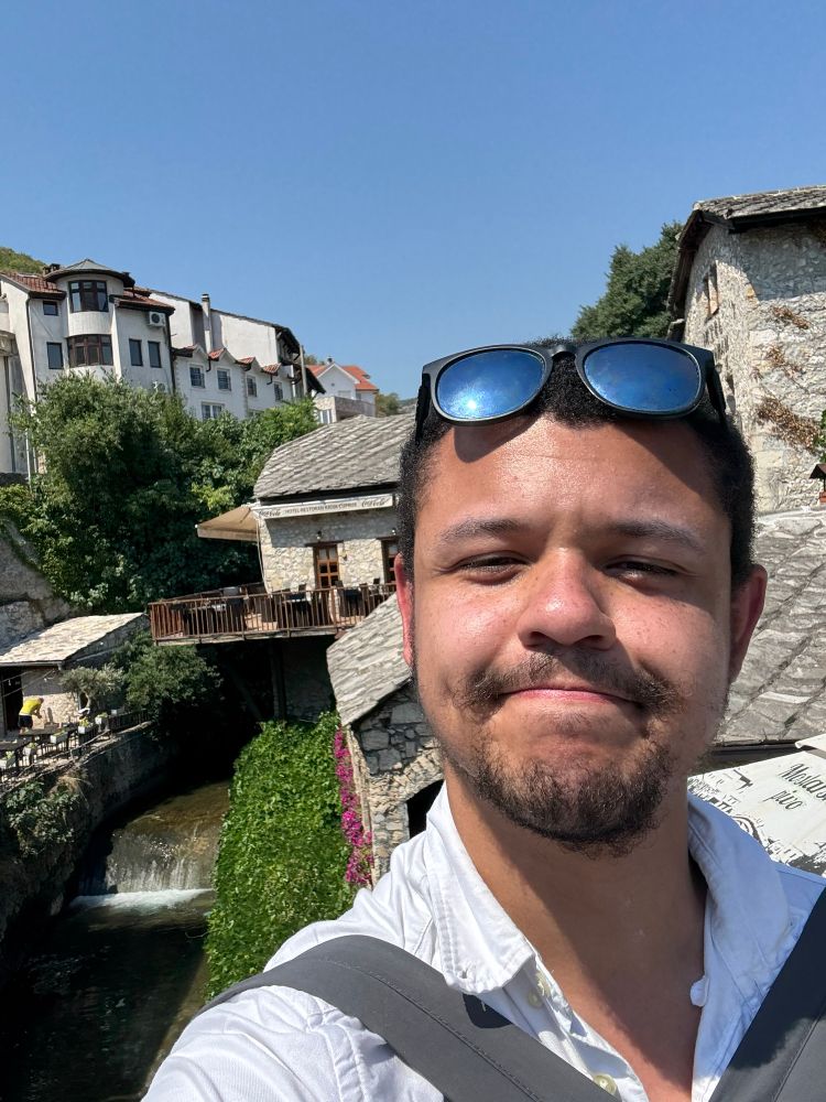 I am smiling in front of a small waterfall in Mostar, Bosnia and Herzegovina, my sunglasses are on my head and my linen shirt has the top two buttons undone