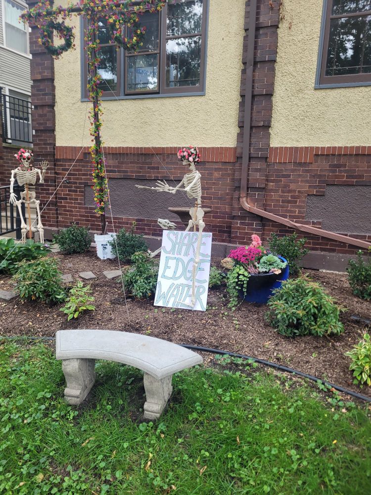 Two yard skeletons in flower crowns next to a handmade sign that says "share Edgewater"