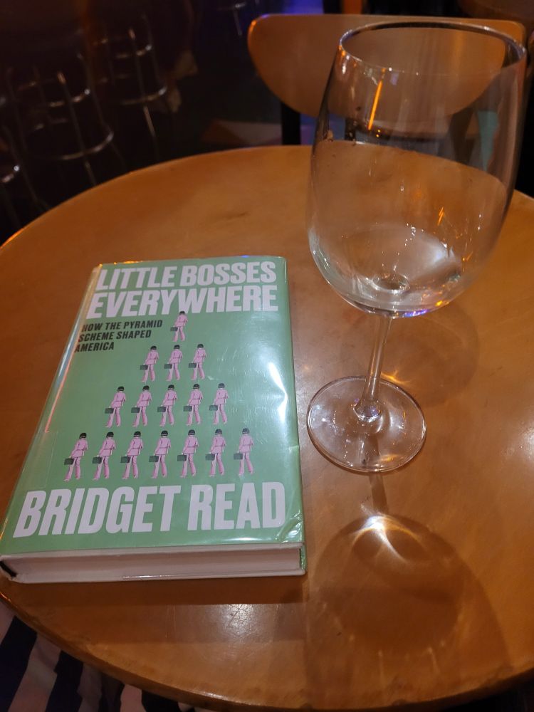 A mostly drank glass of white wine and the book Little Bosses Everywhere: How the Pyramid Scheme Shaped America by Bridget Read on a small round table