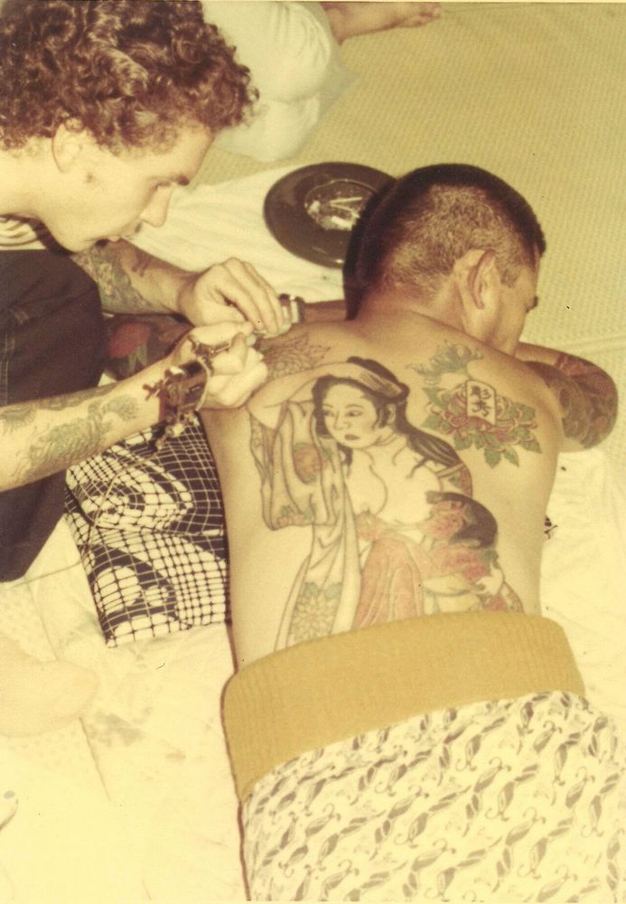 A sepia toned photo showing a young Hardy tattooing a client's back in Gifu, Japan. The tattoo depicts a Japanese woman looking in the distance as her child reaches for milk from her breast.