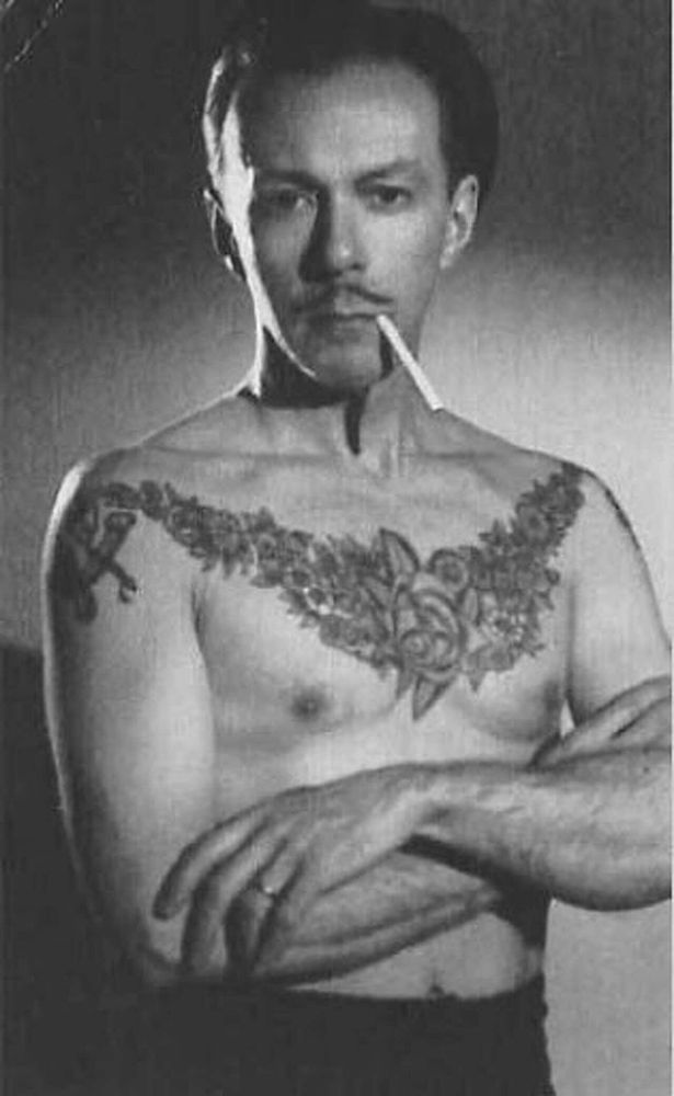 A grayscale portrait photo of Phil Sparrow. His shirt is off, revealing his slender figure and a large floral tattoo spanning from either shoulder across his entire chest. He has an unlighted cigarette in his mouth and his folding his arms.