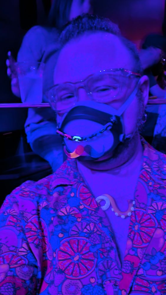 A selfie of me wearing a romper with a fruit print, a tentacle necklace, Stoggles, and a Flo Mask with a mustache mask chain, glowing from a black light.