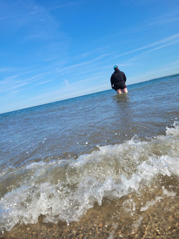 Ich, von hinten fotografiert, stehe bis zum Knie im Meer, im Vordergrund eine kleine, brechende Welle.