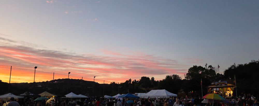 View from the back of the audience field toward the stage (toward the right, appreciating a glowing sunset toward some oak covered hills, over the tops of tents, umbrellas and awnings . The trees in the back are in silhouette with the darkening sky