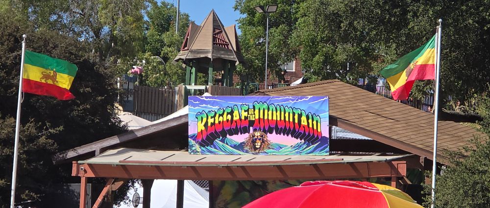 event sign over a wood satge awning with "Reggae on the Mountain" in progressive green, yellow, red. a crowned lion's face floats above a verdant valley that stretches into a pink and blue sky. 2 flag poles topped with Rasta, lion of Judah flags are on either side of the stage.in front of the stage to the right, obstructing view of stage is the top of a red and yellow sun-umbrella. Behind the stage is a wood gazebo and, mostly,  oak trees (Quercus chrysolepis) all across the back of the photo.
