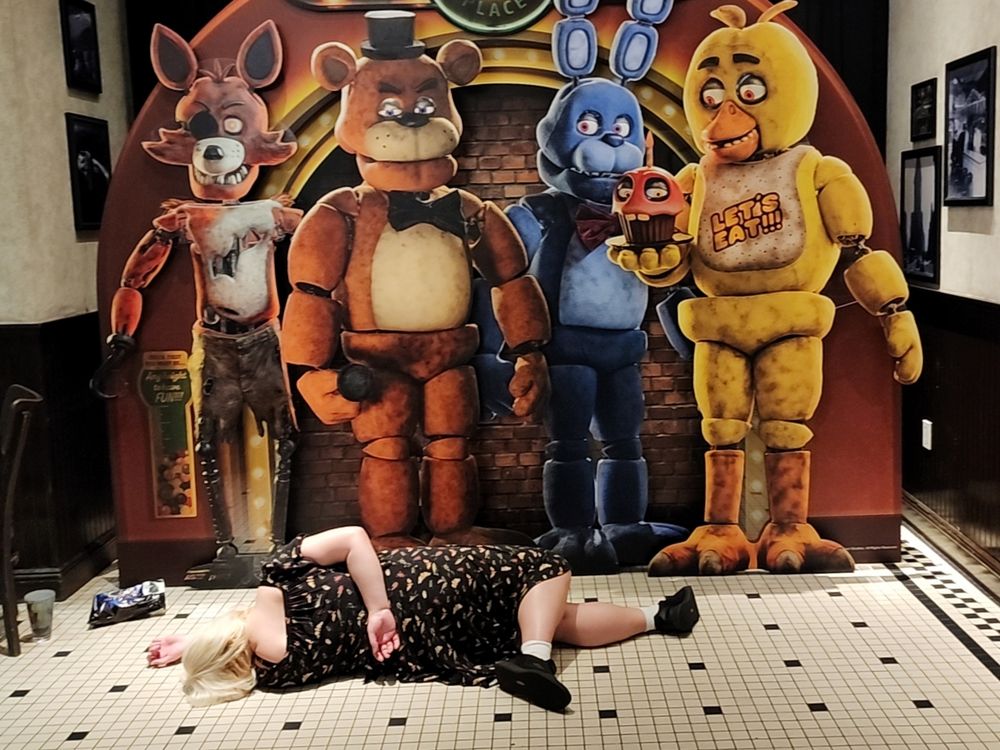 a five nights at Freddy's standee featuring all the animatronics staring down at a blonde woman doing the peter griffin death pose
