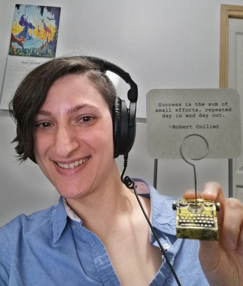A brown-eyed and dark-haired non-binary person with an undercut is displaying the smile of rediscovered motivation.

They are wearing a black noise-reducing headset and they are holding a black and gold card-holder which is shaped like a typewriter.

The card-holder is holding a card on which is printed, in a typewriter-style font, a Robert Collier quote: "Success is the sum of small efforts, repeated day in and day out."