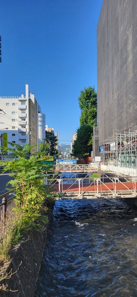 A landscape shot looking over a small river. There are several pedestrian bridges over the river and tall buildings on both sides. In the distant horizon there are mountains.