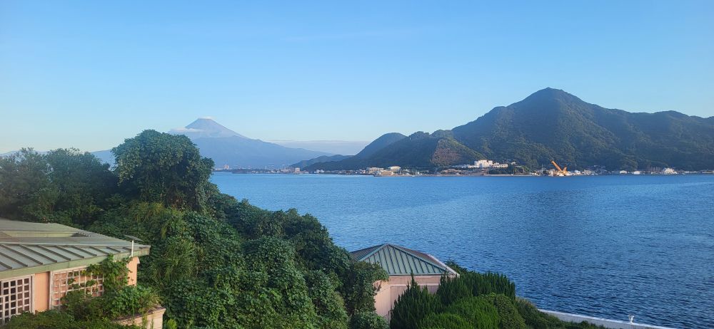 From high up on a balcony, the view overlooks mountainous foliage in the foreground, calm waters behind it, and a small town on the coastline in the distance that sits at the foot of a mountain. Even further in the distance is Mt. Fuji. Its peak is higher than some of the small clouds in front of it.