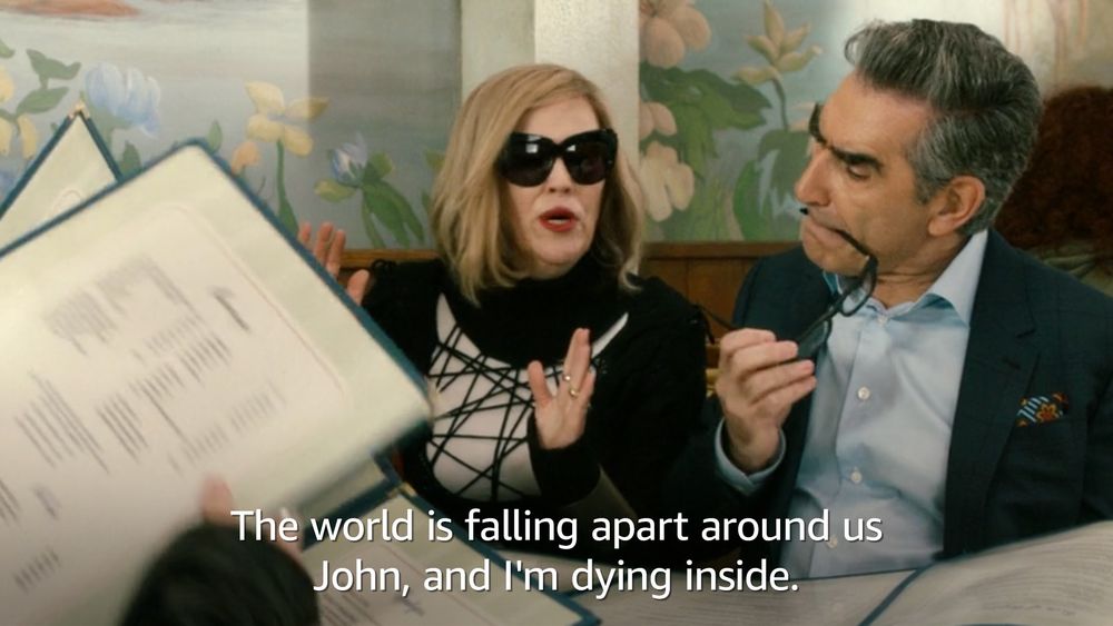 Image still from Schitt's Creek where Moira Rose is wearing a black knit sweater spiderwebbed over a white top. She has black sunglasses on and is seated next to Johnny Rose. David and Alexis are in the background holding their menus out. The dialogue reads, "The world is falling apart around us John, and I'm dying inside." 