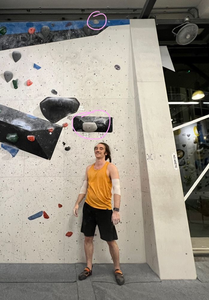 Me, a grinning white dude in a loose orange tank top, standing in front of a climbing wall. The image is edited to have two holds circled in pink showing the v5 I did. The first one is a jug on a small volume, just within my arm's reach from the ground, and then a small hold at the very top of the wall, about a full body+arm reach from the volume the first hold is on.