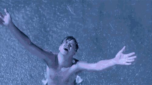 A man standing shirtless in the rain with his arms outstretched, looking up with a triumphant and emotional expression, as rain pours down around him.