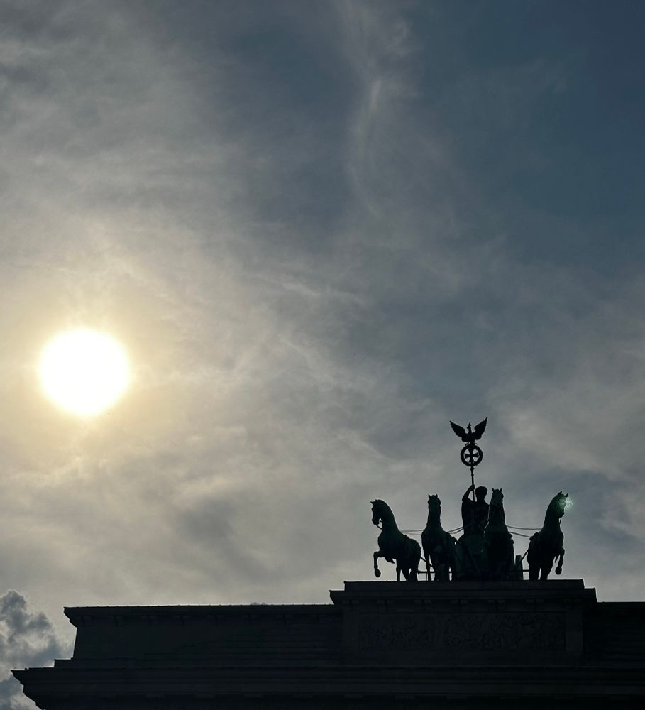 The Brandenburg Gate in Berlin photographed in silhouette, showing the Quadriga statue of a chariot drawn by four horses with the goddess of victory holding a staff and wreath. The sun glows brightly in the cloudy sky to the left, casting the monument into dramatic shadow.