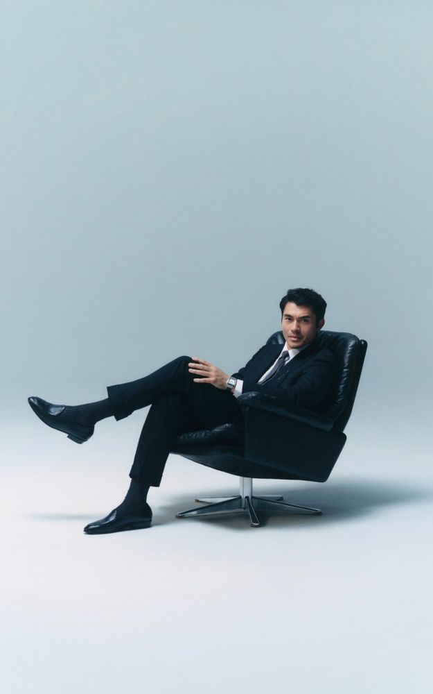 A photo of Henry Golding from a GQ x Cartier photoshoot. He's sat cross-legged in a suit on a mid-century style chair.