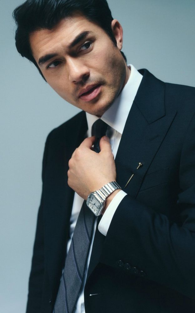 A photo of Henry Golding from a GQ x Cartier photoshoot. He's wearing a suit and is adjusting his tie.