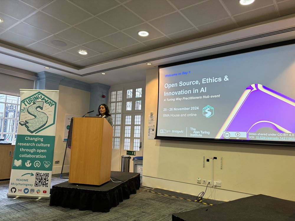 A speaker - Malvika, an Indian woman - stands at a podium opening a conference titled "Open Source, Ethics & Innovation in AI." The event is organized by The Turing Way and there is a banner advertising the project.