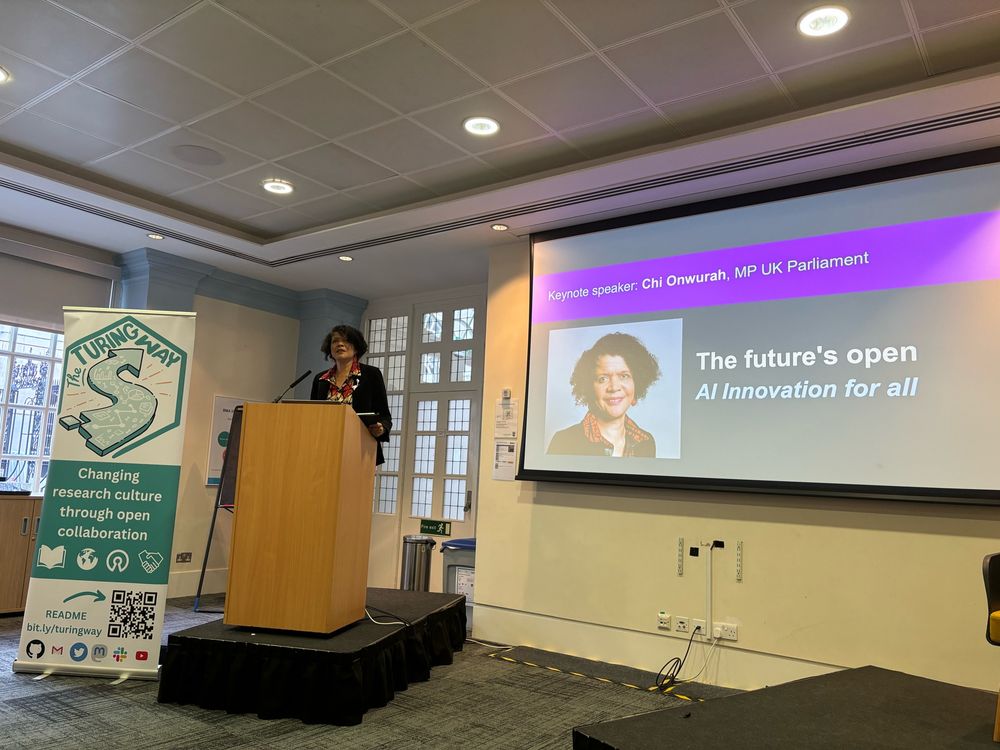 A black woman (Onwurah) stands at a podium with a banner for "The Turing Way" beside them. A slide is projected, featuring the keynote speaker's name and the theme "The future's open - AI Innovation for all."