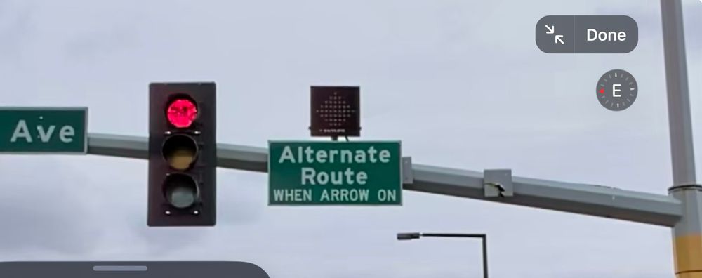 A traffic light support extending over a road. The light is red. To the right of the traffic light is a sign that says, "Alternate route when arrow on." Above that is an unlit black box.