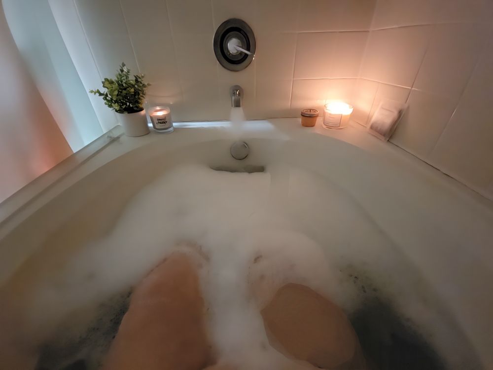 An oversized bathtub with lit candles and faux greenery, filled with water and bubbles. A woman's thighs peek out from beneath the bubbles.