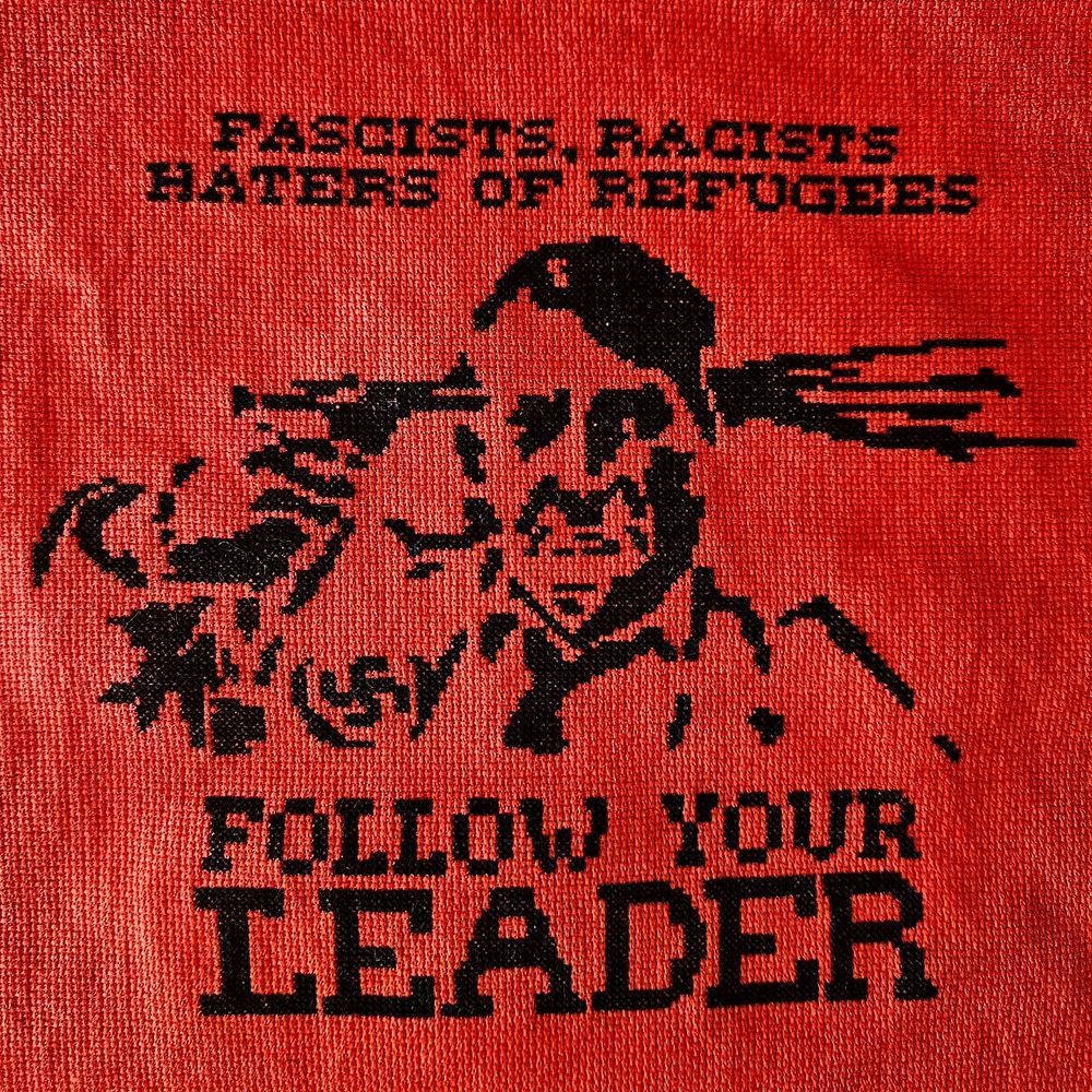 cross stitched design of hitler holding pistol to his head with his right hand and a spray of blood on the left side. text above the design reads "fascists, racists, haters of refugees" and the text below the design reads "follow your leader"