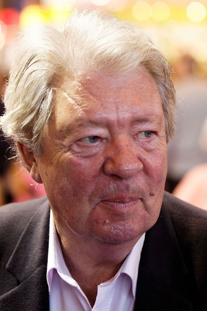 Portrait de Jean-Jacques Sempé, il a des cheveux blancs et bien coiffés. Il porte une chemise blanche et un blazer noir élégant. L’arrière-plan est flou, avec des lumières jaunes diffuses, suggérant un environnement intérieur éclairé.