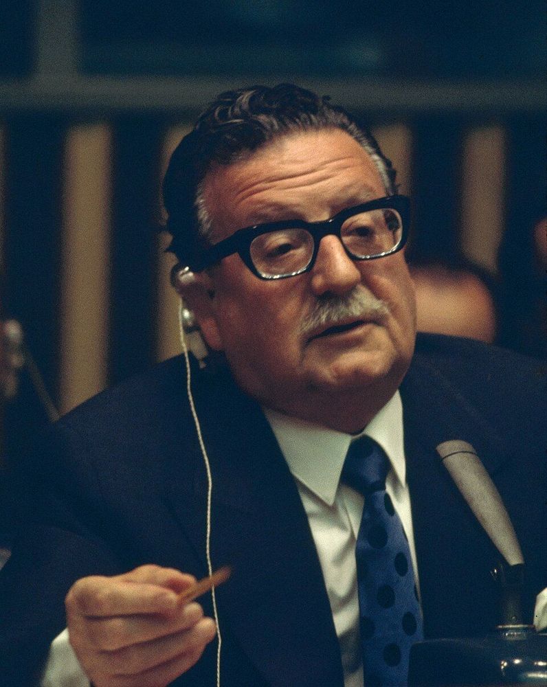 Salvador Allende, vêtu d'un costume sombre et d'une cravate à pois bleus et blancs, est assis à une table lors d'une réunion ou conférence. Il tient un stylo dans sa main droite et porte un casque audio, avec un microphone placé devant lui
