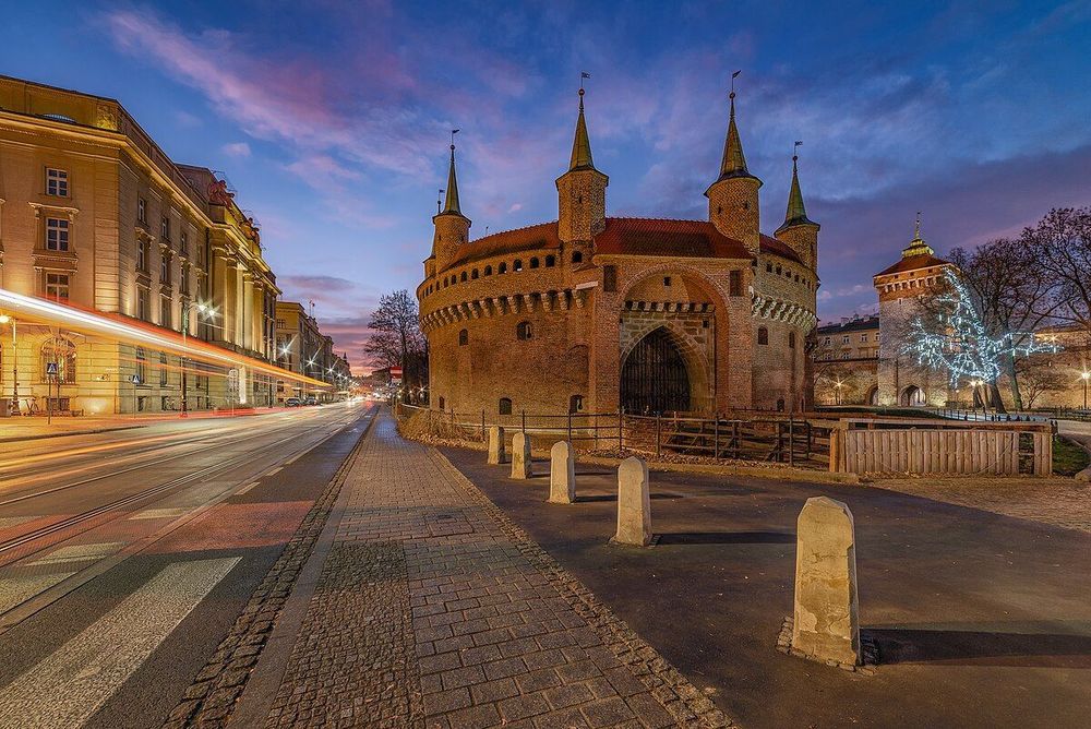Vue de la Barbacane de Cracovie avant le lever du soleil. Le ciel est teinté de rose et de violet. Le bâtiment en briques rouges, de forme circulaire avec des tourelles, est éclairé, tout comme la rue pavée et les bâtiments environnants. On peut voir les traces lumineuses des phares de voitures sur la route de gauche.