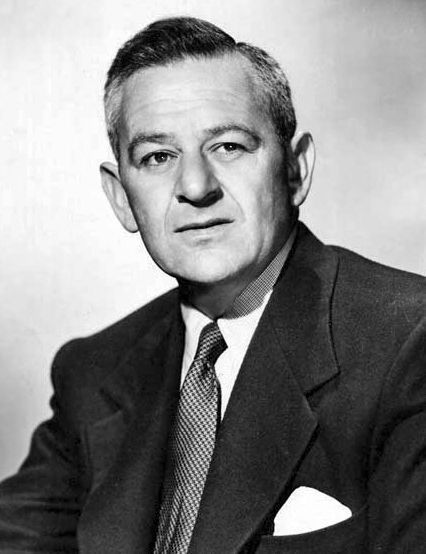 Photo en noir et blanc de William Wyler, vêtu d’un costume sombre et d’une cravate. Il a un mouchoir blanc soigneusement plié dans la poche de sa veste. Le fond de l’image est uni et clair, mettant en valeur la silhouette de Wyler