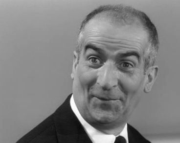 Photographie en noir et blanc de l'acteur Louis de Funès, en train de faire une grimace. Il est vêtu d’un costume sombre avec une chemise blanche visible au niveau du col. L’arrière-plan est uniforme et gris clair.