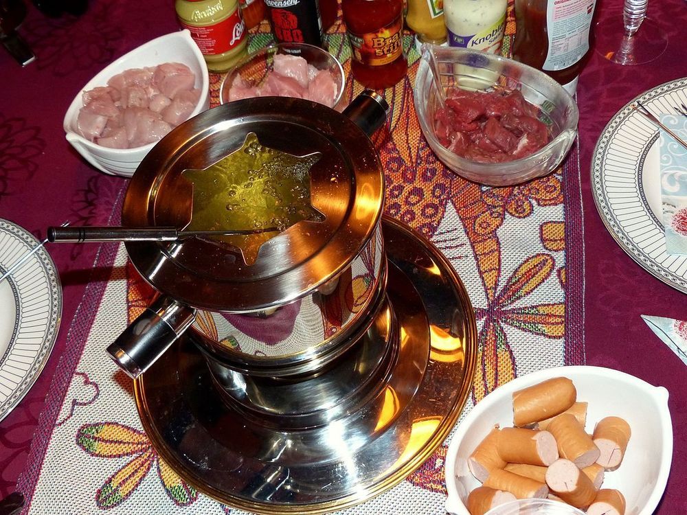 Une table conviviale dressée pour une fondue de la Saint-Sylvestre. Au centre, un caquelon contenant du bouillon frémissant ou de l’huile chaude, entouré d’assiettes garnies de morceaux de viande prêts à être cuits. Des piques à fondue reposent sur le bord du récipient, témoignant d’un moment de partage festif.