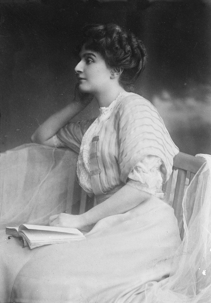 Image en noir et blanc de Marie Bonaparte, assise de profil sur une chaise en bois. Elle porte une robe à manches bouffantes et un corsage orné. Ses cheveux sont coiffés en chignon. Elle tient un livre ouvert sur ses genoux, suggérant qu’elle est en train de lire.