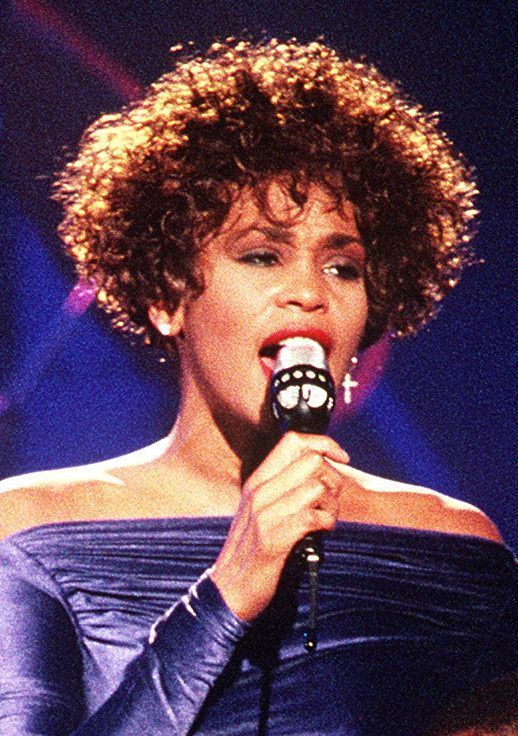 Photo de Whitney Houston, cheveux frisés, tenant un microphone, vêtue d’une robe bleue et avec des cheveux bouclés. L’arrière-plan est sombre avec un peu de lumière, indiquant probablement un cadre de scène ou de performance.