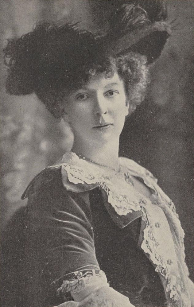 Portrait en noir et blanc de Cecile Chaminade, vêtue d’une robe élégante avec des détails en dentelle, portant un chapeau orné.  L’arrière-plan est flou et indistinct, mettant l’accent sur la personne.